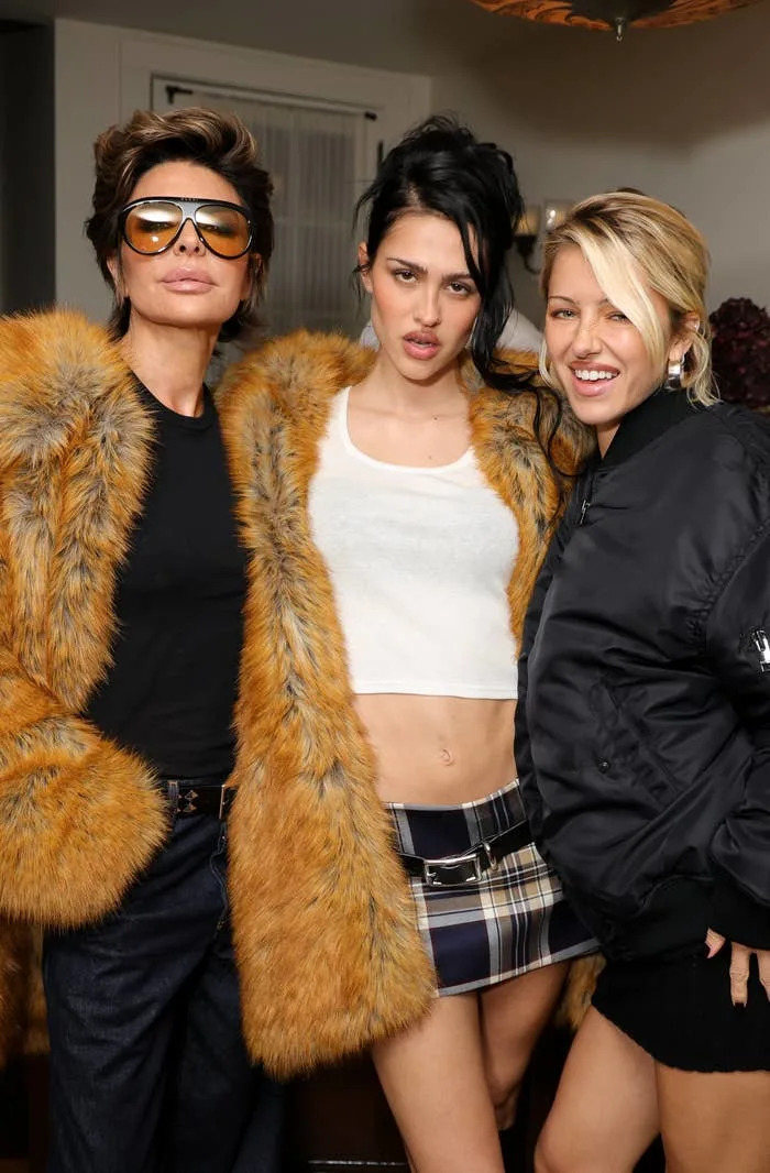 Three people pose at an event: one in a faux fur coat with sunglasses, another in a crop top and plaid skirt, and the third in a bomber jacket
