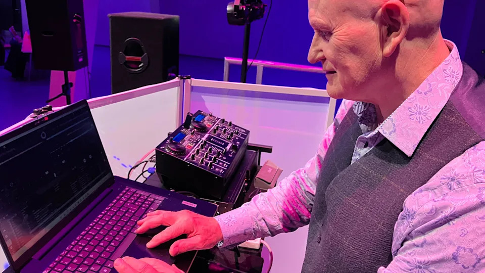 A man who looks like he is in his 70s dressed smartly in a floral shirt and grey waistcoat, he has a bald head and is DJing on a laptop. 