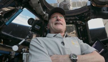 A person wearing glasses, a cap, and a light gray shirt stands inside a spacecraft cockpit with visible controls, monitors, and a window showing the sky and parts of the space station outside. The person is looking upward and smiling.