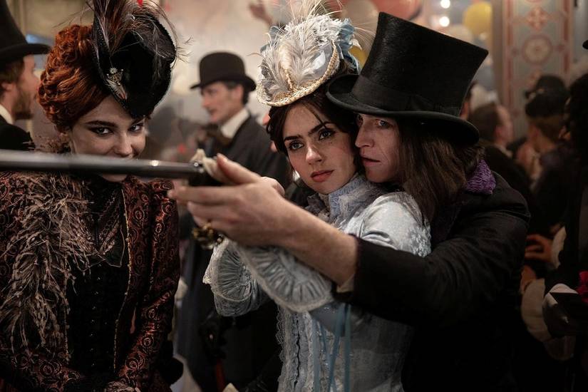 In a scene from Luc Besson's Dracula, Dracula (Caleb Landry Jones) helps Mina (Zoë Bleu) aim a gun at a carnival game, while he wears a top hat that looks a bit like the Gary Oldman version of Dracula in Francis Ford Coppola's movie.