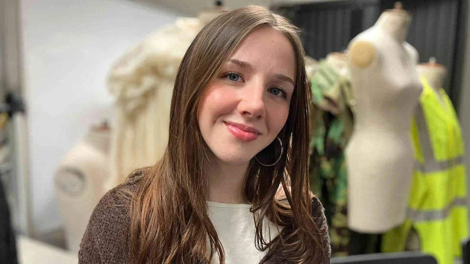 Scarlett Baker has long brown hair and is wearing a textured brown cardigan over a light top. Behind her, there are some mannequins, one features a cream‑coloured dress, another has a green patterned outfit, and a third is dressed in a bright yellow high‑visibility jacket but they are not in focus. 