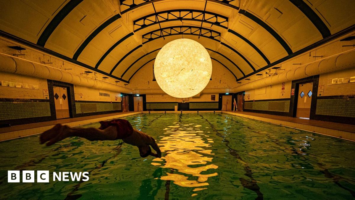 A man dives into a dimly lit swimming pool. Above the pool, suspending from an arched ceiling is a large glowing sun.