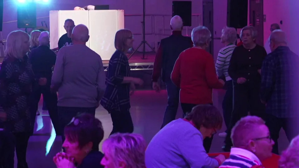 A group of older people dancing at a disco. There are pink and blue disco lights. 