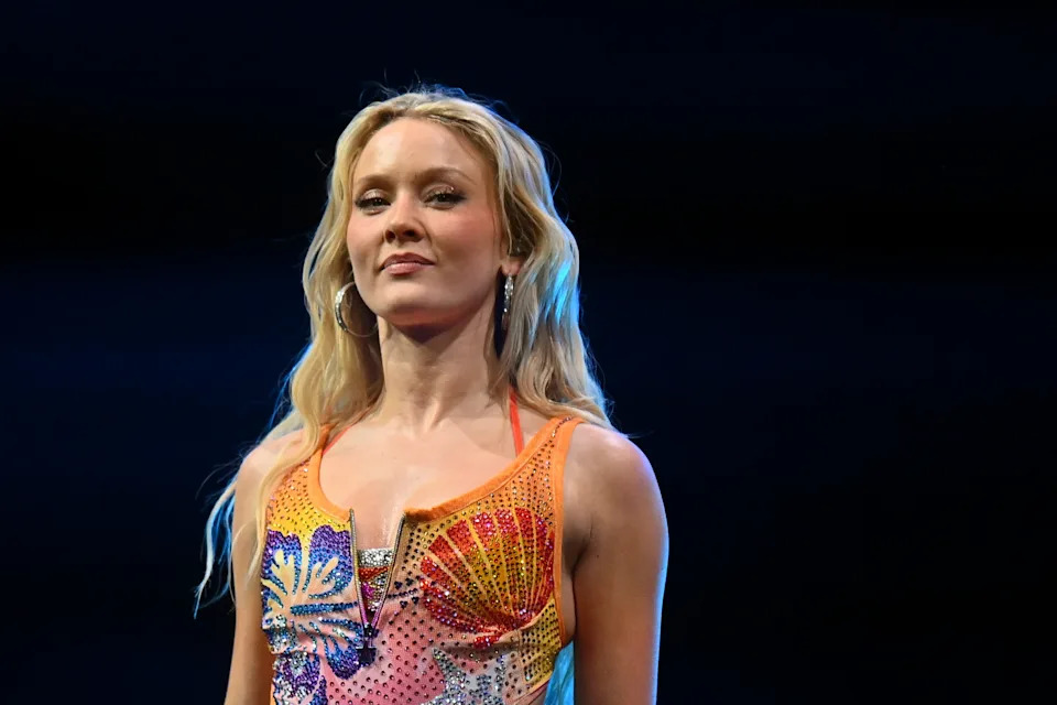 Zara Larsson in a vibrant, patterned dress with wavy hair stands on stage, looking confidently ahead, possibly at a celebrity event