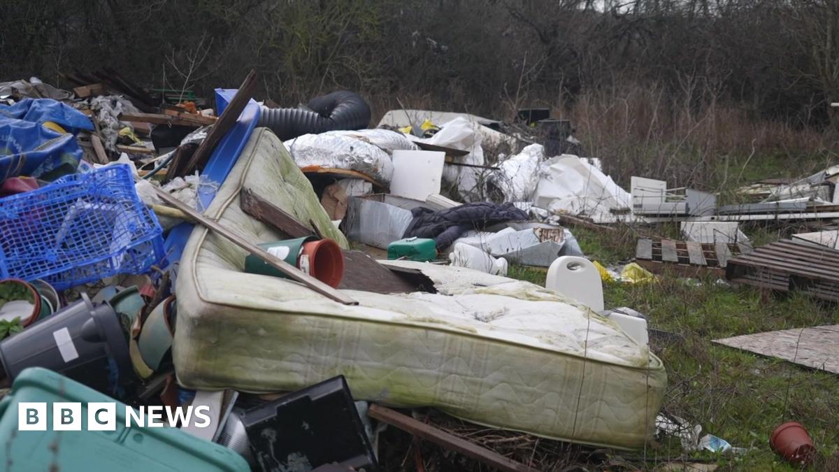 A large pile of fly‑tipped rubbish in a grassy area, including an old stained mattress, plastic crates, broken furniture and assorted household waste.