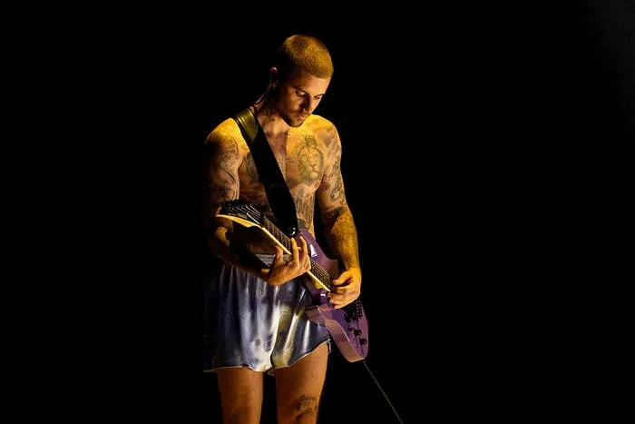 A tattooed person plays an electric guitar on stage, wearing a sleeveless top and shorts