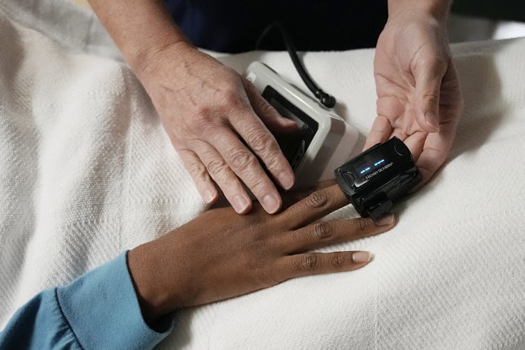 A black woman has a pulse oximeter attached to her fingers by a white nurse.