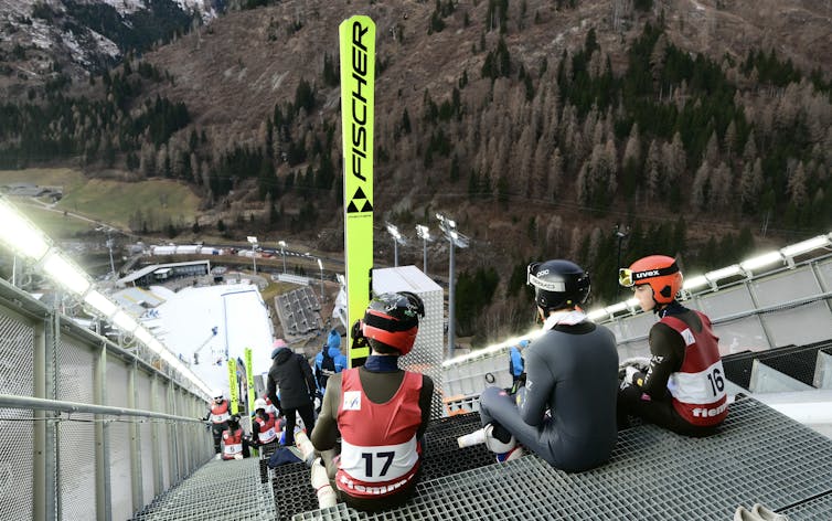 Three skiers sit at the top of a ski jump. Their view shows how much dry, snow-free ground is around the jump area