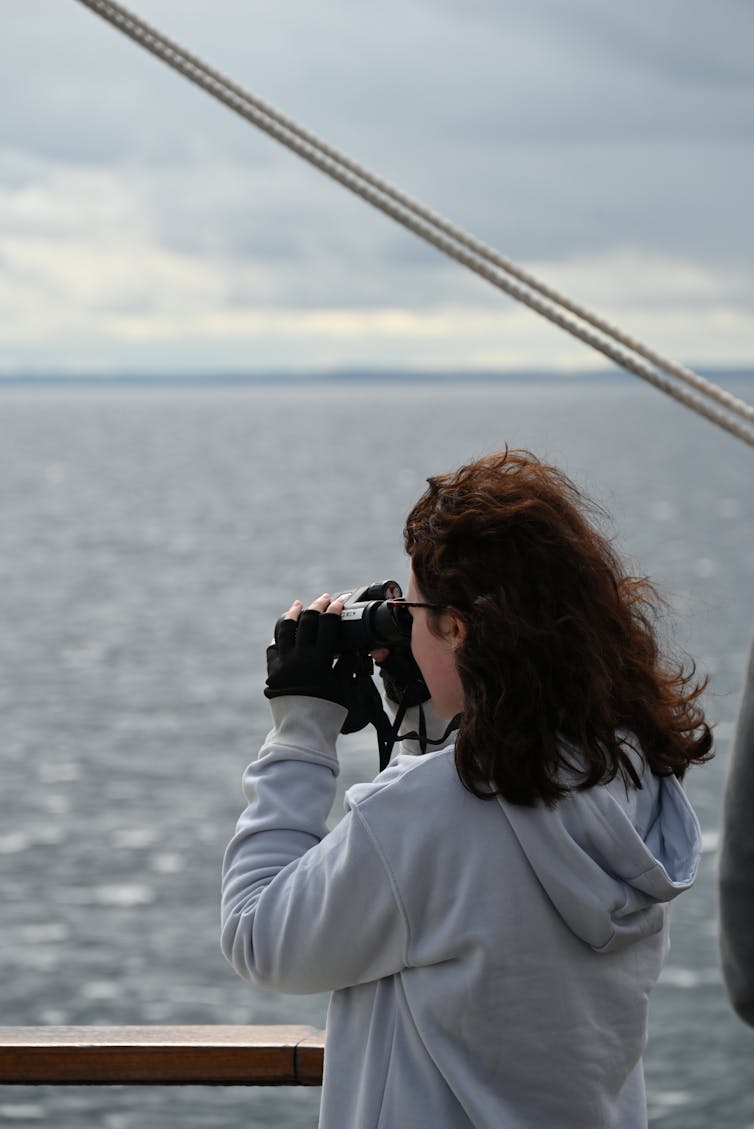 young woman with long brown hair holds binoculars and gazes out to sea