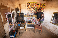 A man and child stand in a small barber shop next to an electric hair razor and phone charging station