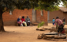 Two people interview an elderly man sitting on a bench outside his house