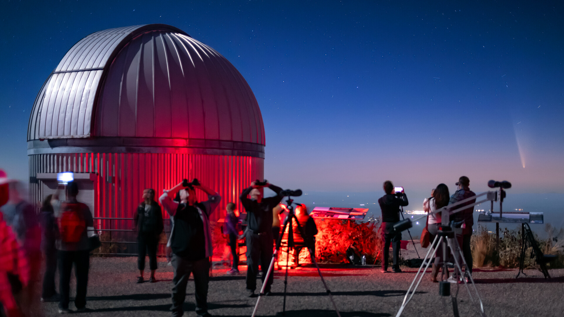 A circular dome of a telescope is illuminated in red light as a crowd of people holding binoculars and telescopes look at a pink and blue night sky.