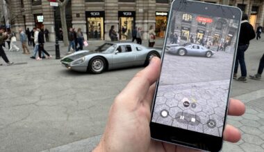 A smartphone is held up to shoot a video of a classic sports car on a street.