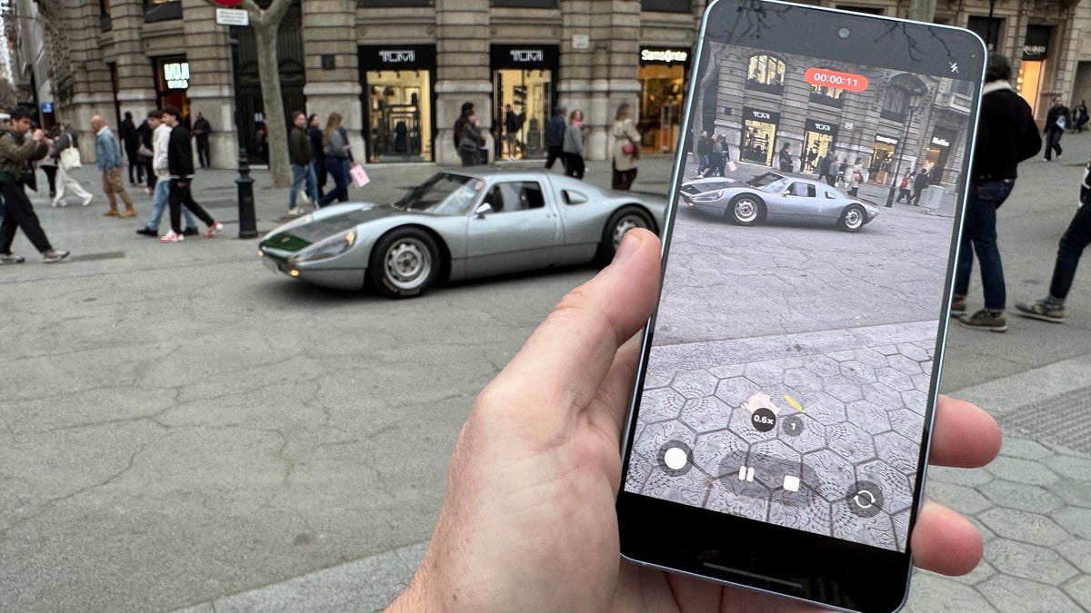 A smartphone is held up to shoot a video of a classic sports car on a street.