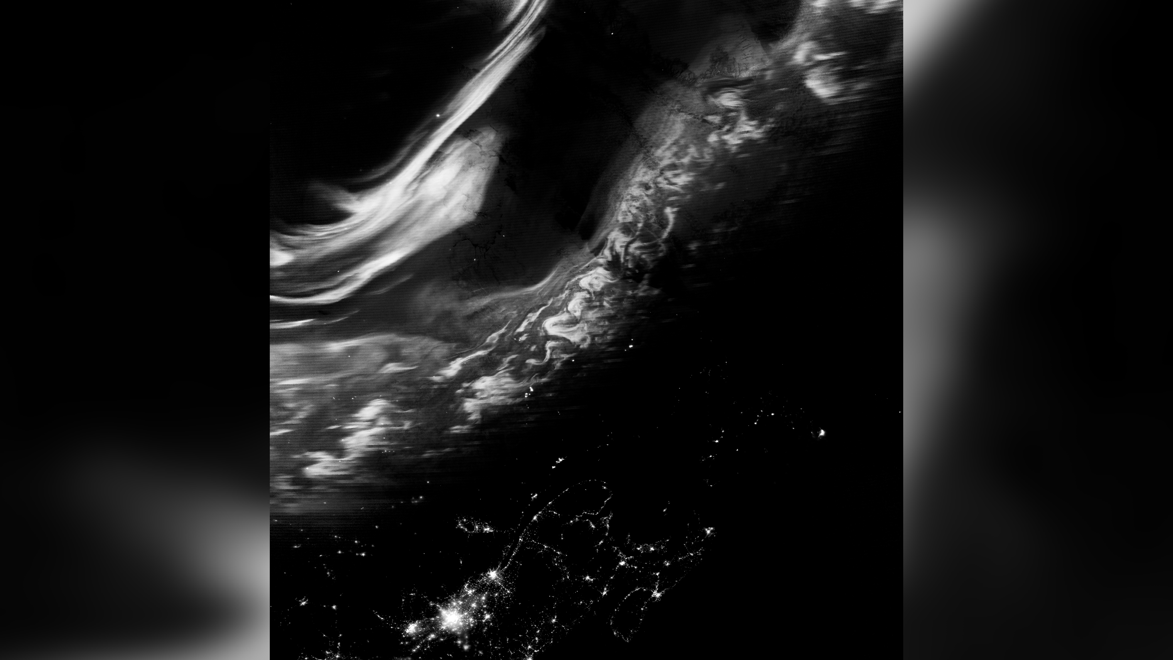 grayscale image showing bright sweeping structures - auroras, shining bright over specks of city lights below.