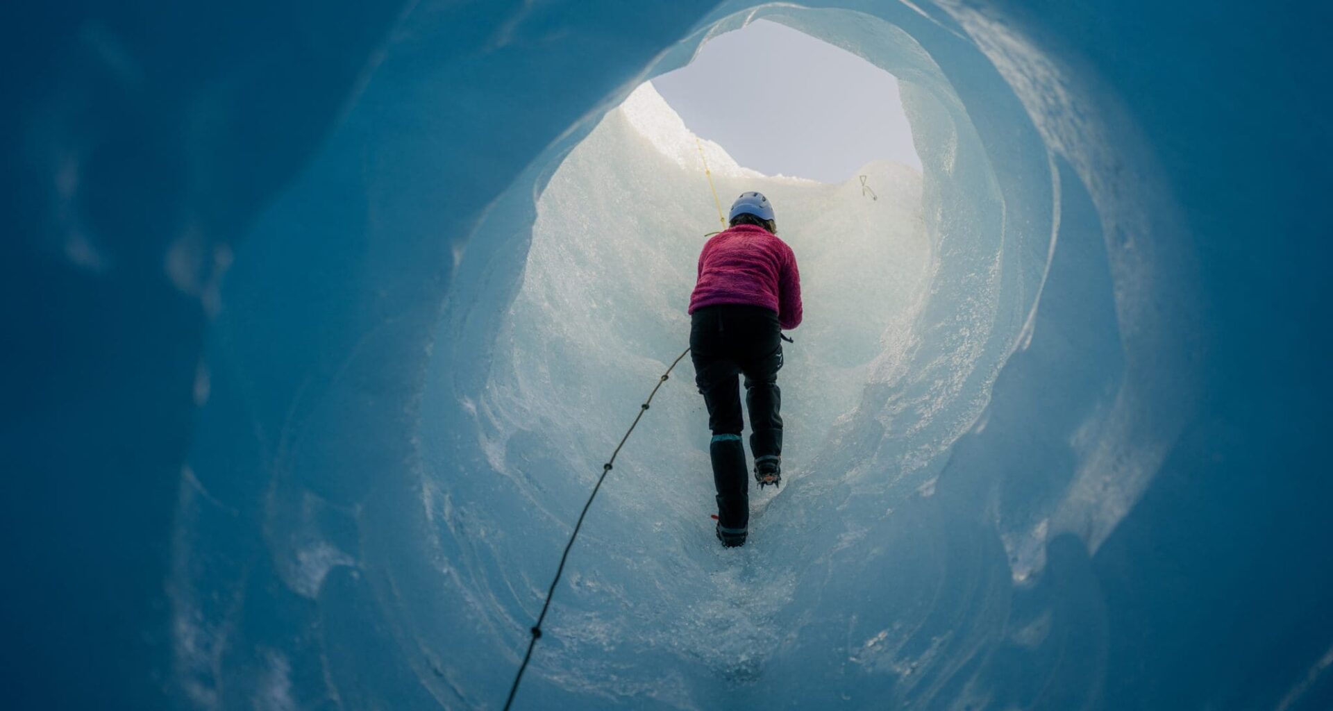 Scientist wearing crampons is climbing a rope while ascending through a stunning deep blue ice cave within a massive glacier