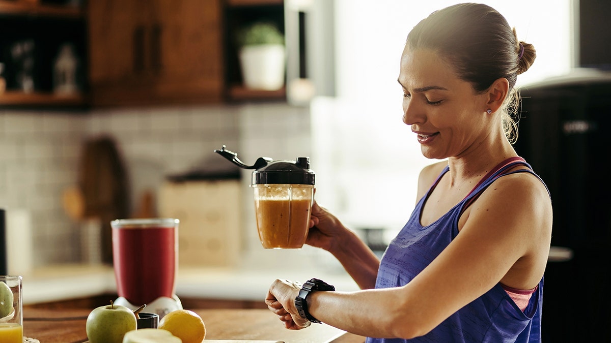 Woman in workout clothes drinking healthy smoothie and checking the time on her wristwatch in the kitchen, indicating she is intermittent fasting.