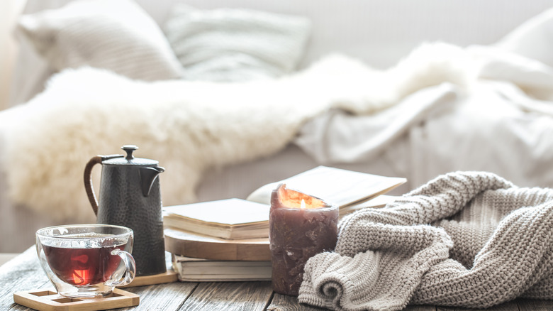 A calming setting with a cup of tea, tea kettle, lit candle, and book, with pillows and white fluffy blankets in the background