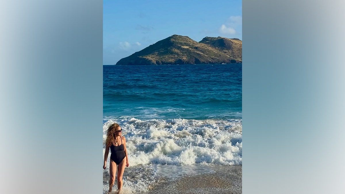 Jennifer Grey walks through the surf in a black one-piece swimsuit with waves crashing behind her.