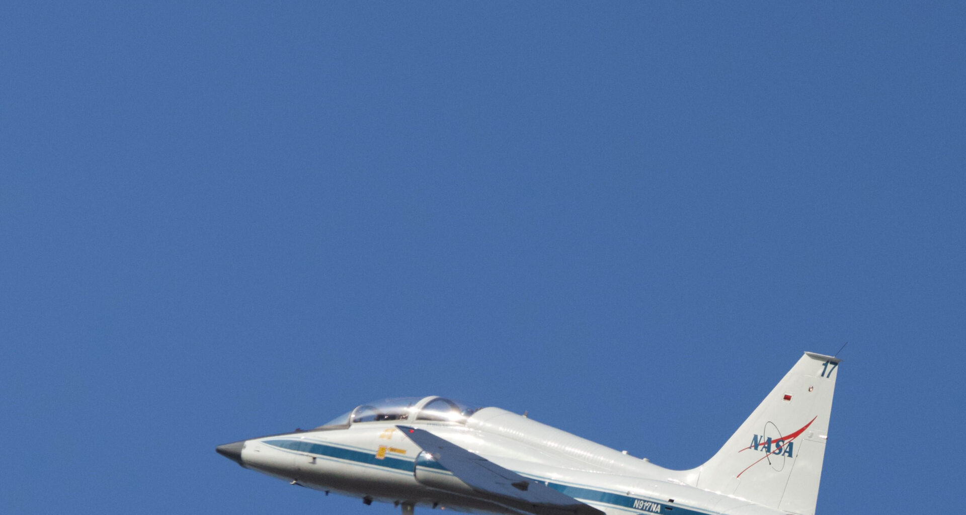 A white plane rises upward to the left against a blue sky. At top left, the waning crescent moon is faintly visible. The plane has a blue stripe low on its body and a version of the NASA insignia on the rudder.