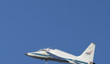 A white plane rises upward to the left against a blue sky. At top left, the waning crescent moon is faintly visible. The plane has a blue stripe low on its body and a version of the NASA insignia on the rudder.
