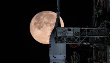 A white rocket nose with the circular NASA logo stands next to the metal gantry underneath a glowing large full moon in a black night sky