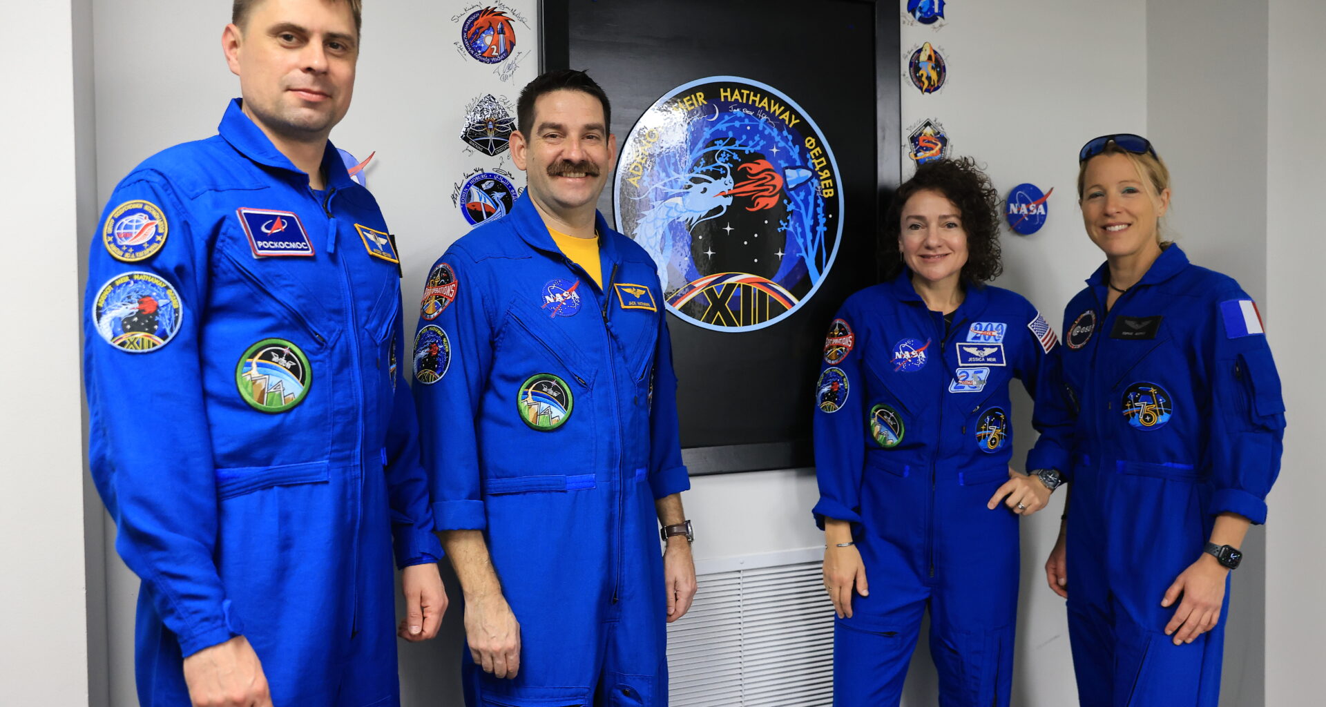 Four people - two men on the left and two women on the right - pose with the Crew-12 mission insignia. They are all wearing blue jumpsuits with various patches on them. The insignia is on the wall, framed in a black recess. Autographed patches are stuck on the wall around the black frame.