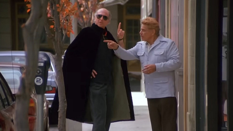 Frank Costanza wears a white coat while talking to a mysterious bald man wearing a cape and sunglasses in 