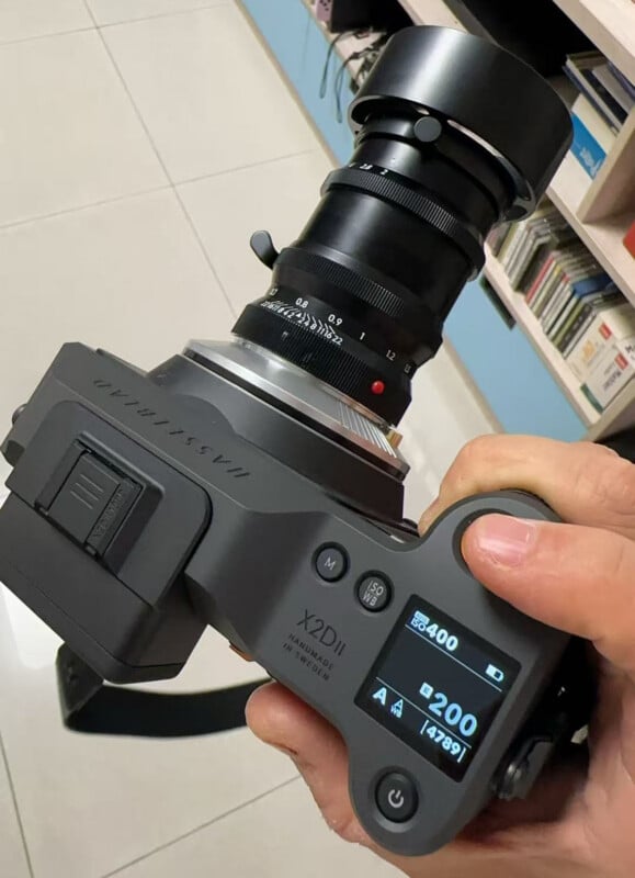 A hand holds a Hasselblad X2D II camera with a lens attached. The camera's display shows settings including ISO 400 and shutter speed 1/200. Bookshelves are visible in the background.