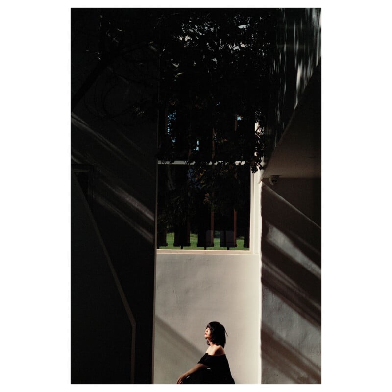 A person sits alone in a sunlit corner, surrounded by deep shadows and dappled light streaming through a window, creating a dramatic and contemplative atmosphere.