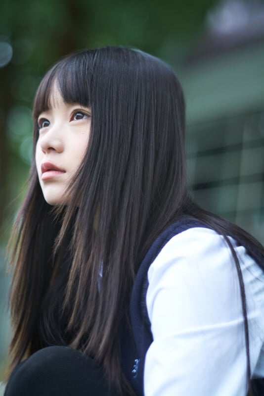 A young person with long straight black hair and bangs, wearing a white shirt and dark vest, looks thoughtfully into the distance with a blurred outdoor background.