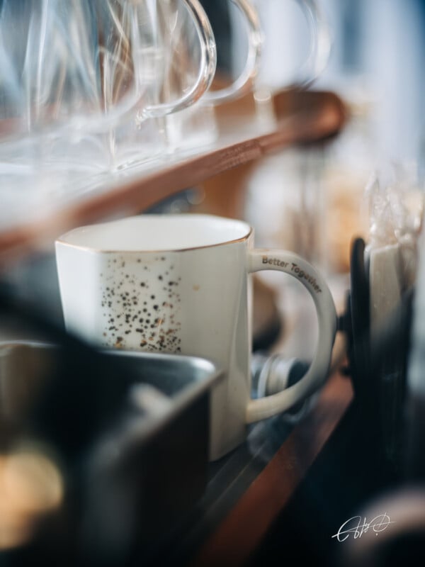 A white ceramic mug with black speckles and the words "Better Together" on the handle sits on a shelf among glassware and kitchen items.