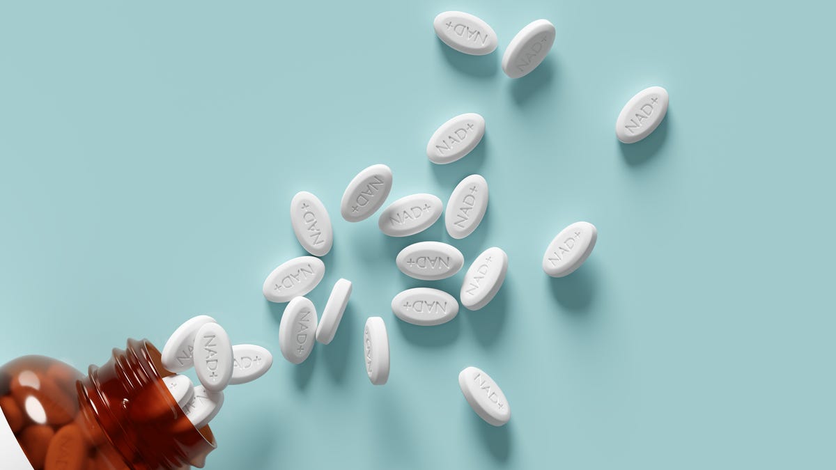 White pills labelled NAD+ spilling out of a brown pill bottle onto a light blue surface.