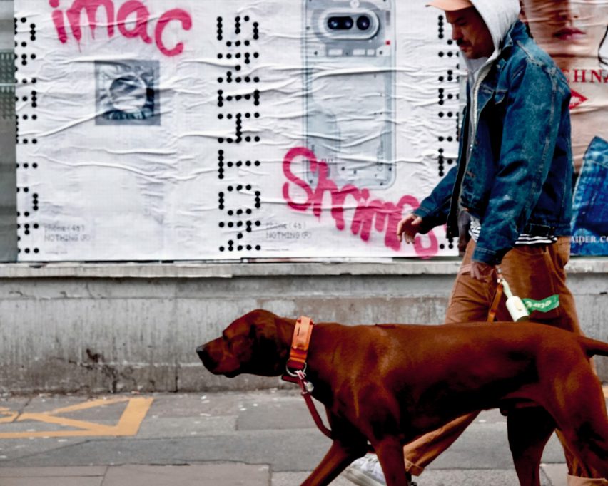 Photo of a street scene with a person walking their dog in the foreground and an advertising posted for a phone in the background grafittied in hot pink with the words iMac and shrimps