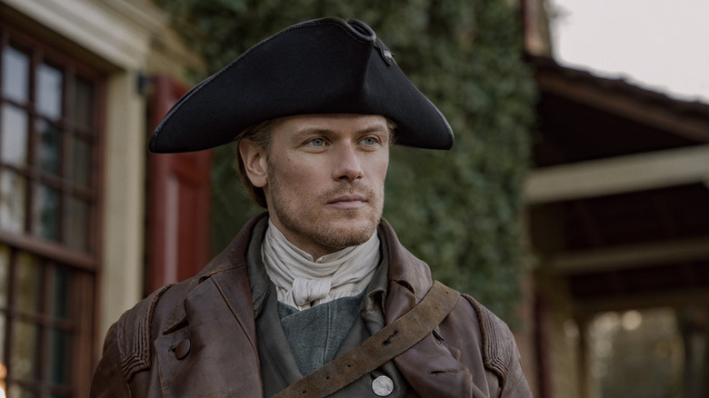 Jamie Fraser (Sam Heughan) stands in a street looking ahead somberly in Outlander