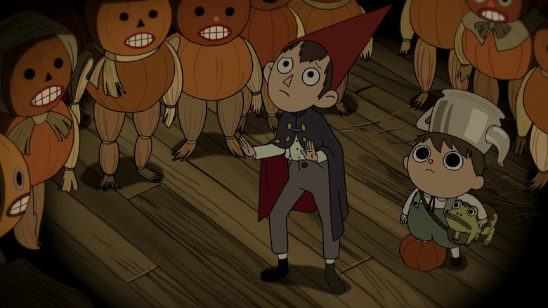 Wirt holds his hands up defensively as Greg stands beside him holding a frog under his arm while the two are surrounded by blank faced Pumpkin People in 