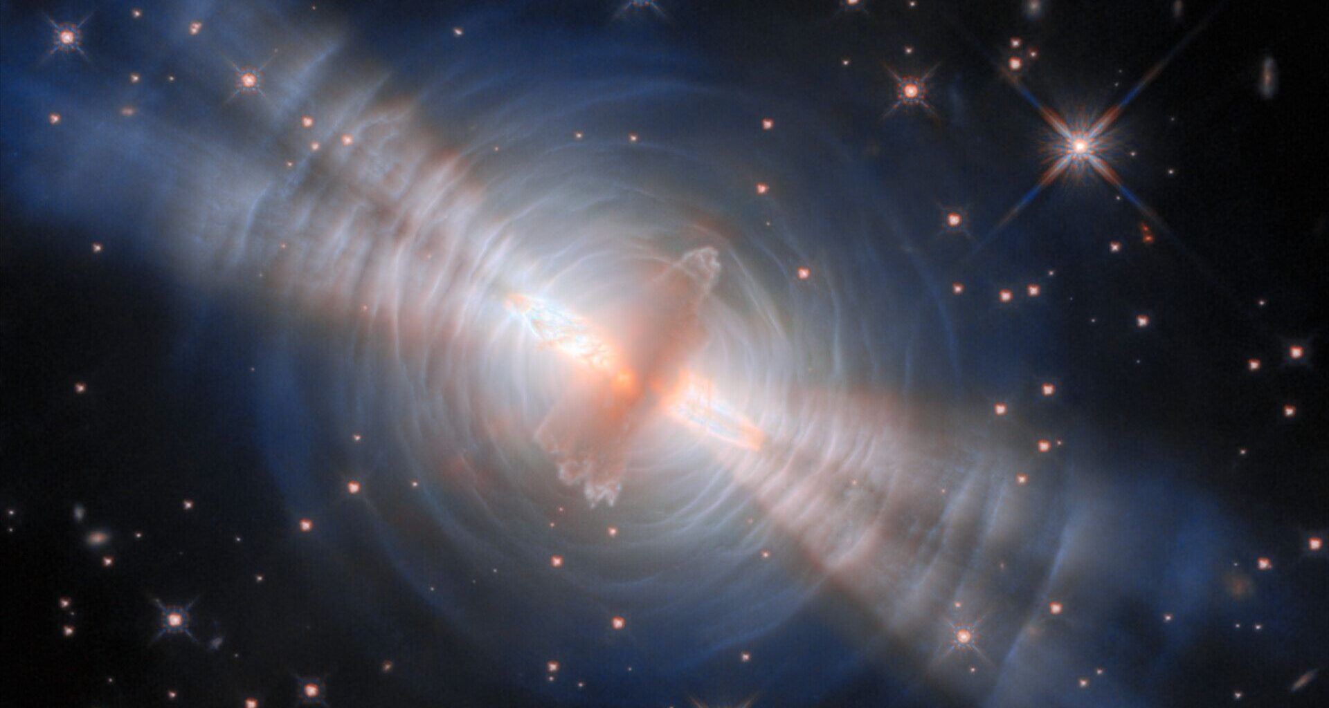 An image of the egg nebula, with a glowing streak of pink gas in the middle surrounded by concentric circles of white like with four diagonal beams of light streaking from top left to bottom right all in front of a deep space starry background