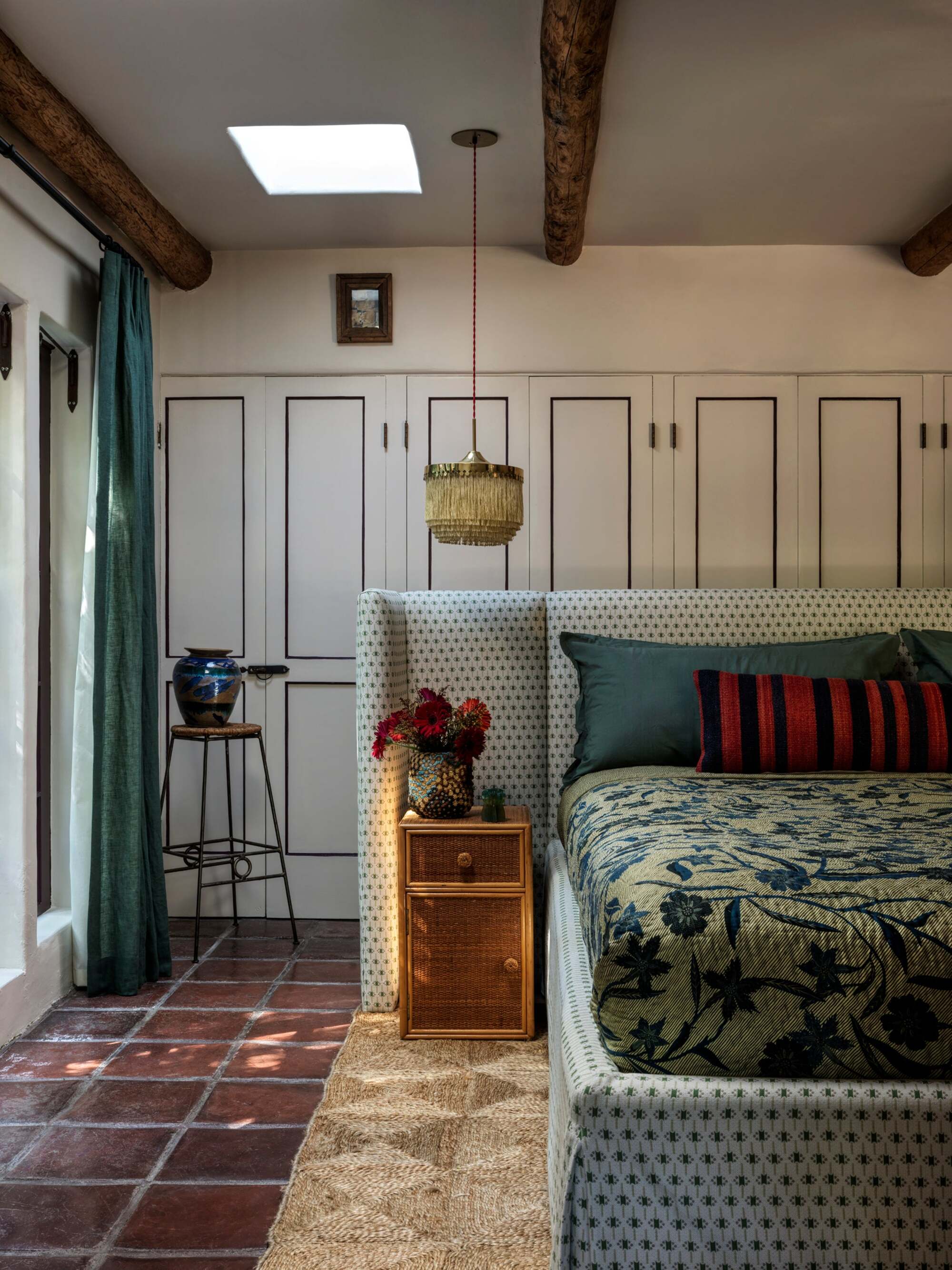 bedroom with floating bed with upholstered bedhead, rattan side tables, tapestry-style bedcover, cabinets behind, stool with vase on top, blue curtain with pendant light hanging down