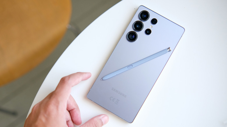 The Samsung Galaxy S25 Ultra, and its stylus, on a white table, with a hand reaching out to grab it.