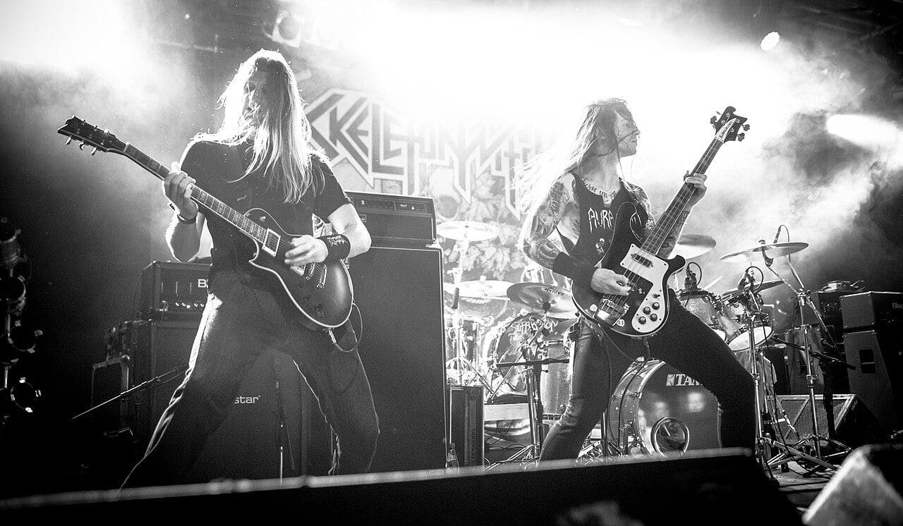 Black-and-white photo of two long-haired guitarists performing on stage with a drummer in the background, surrounded by amplifiers, bright backlighting, and stage smoke during a live metal concert.