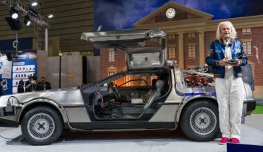A man with white hair, wearing a blue jacket and white pants, stands next to a DeLorean car with gull-wing doors open, in front of a backdrop resembling the "Back to the Future" clock tower.