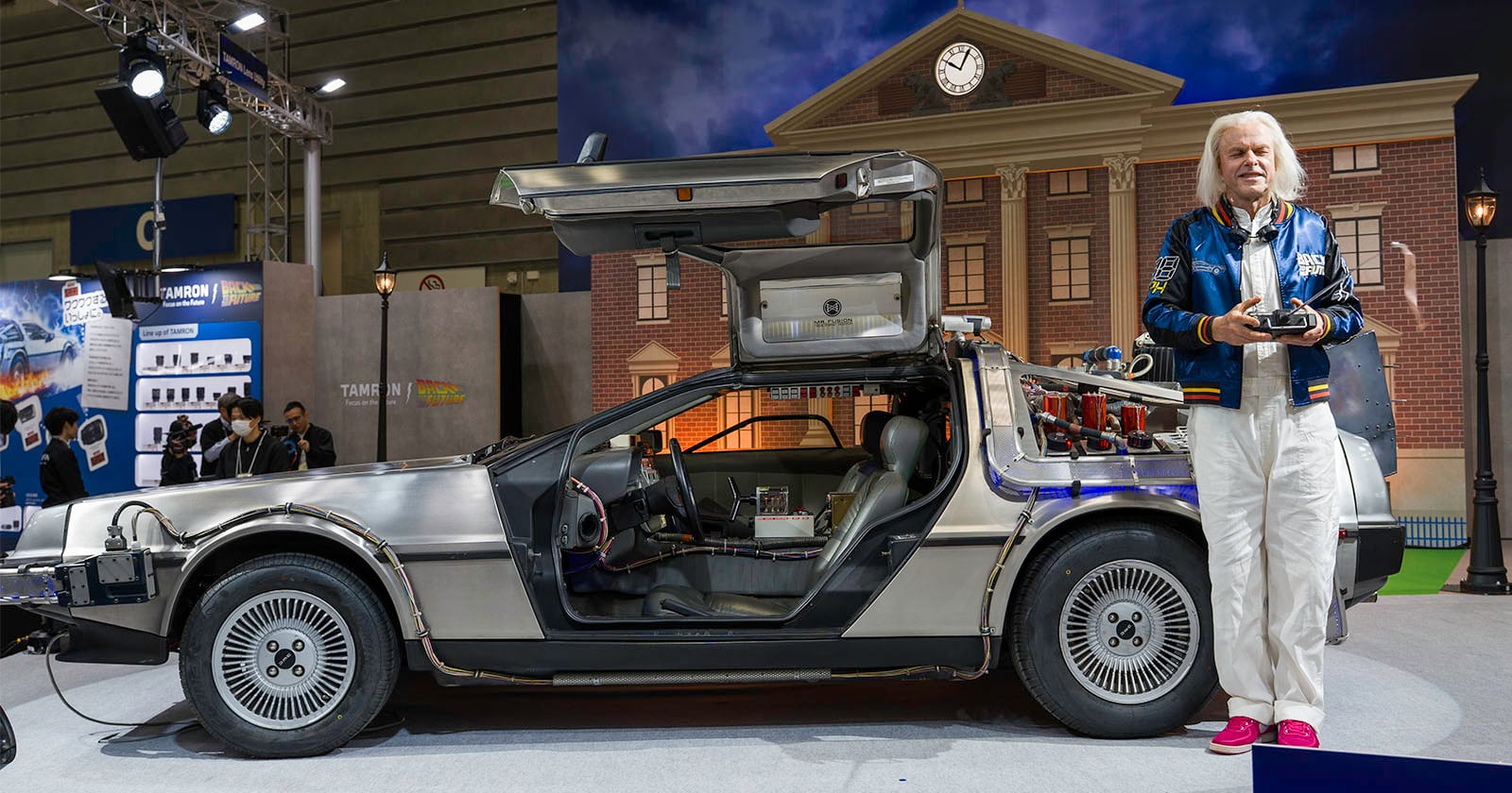 A man with white hair, wearing a blue jacket and white pants, stands next to a DeLorean car with gull-wing doors open, in front of a backdrop resembling the "Back to the Future" clock tower.