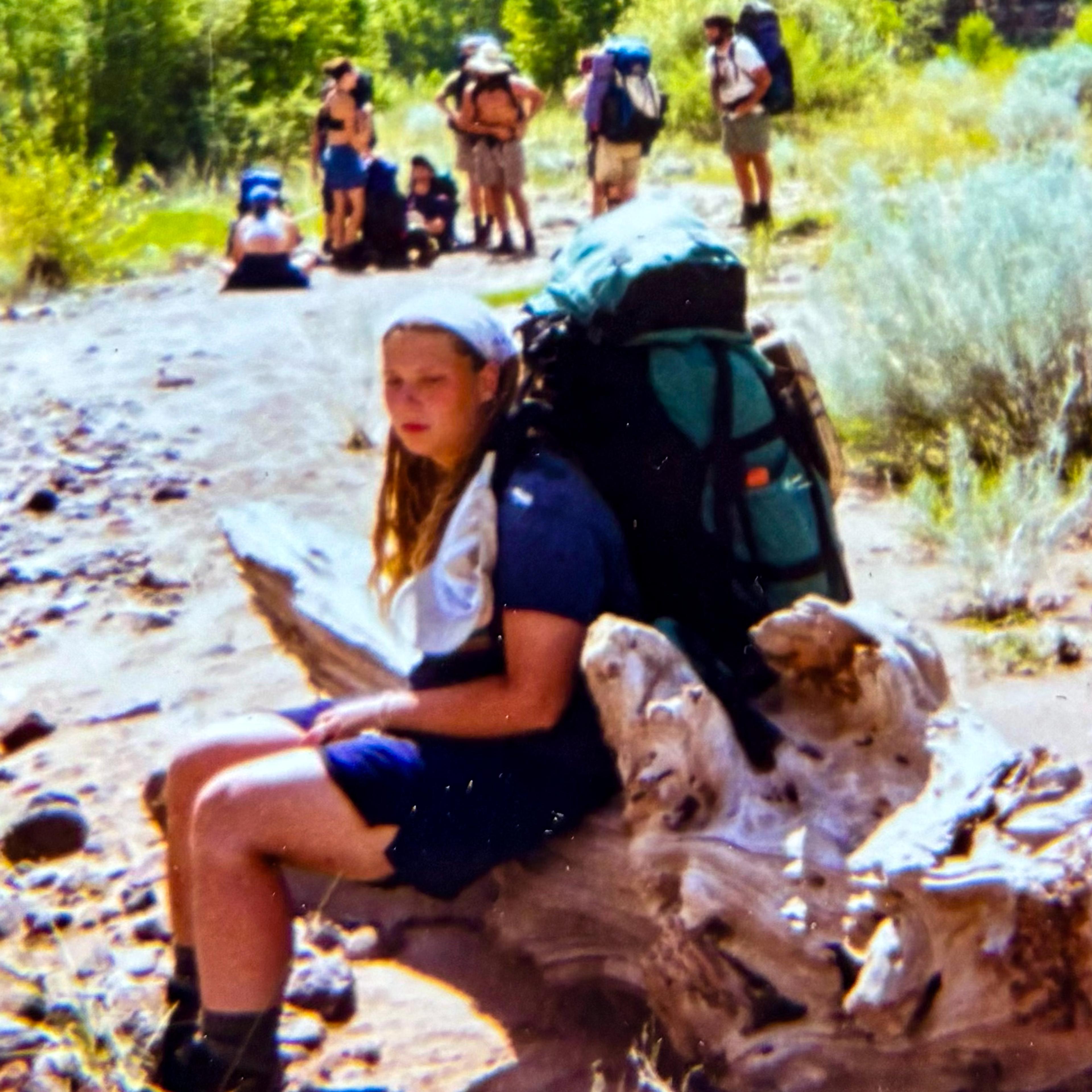 A person with a backpack resting on a log by a rocky riverbank with a group of people in the background.