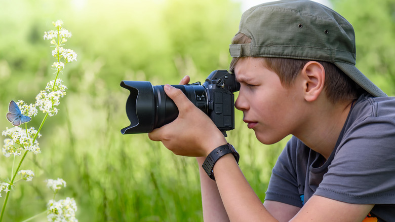 A teen in a backwards baseball cap taking a photo of a butterfly in a field with a large black camera