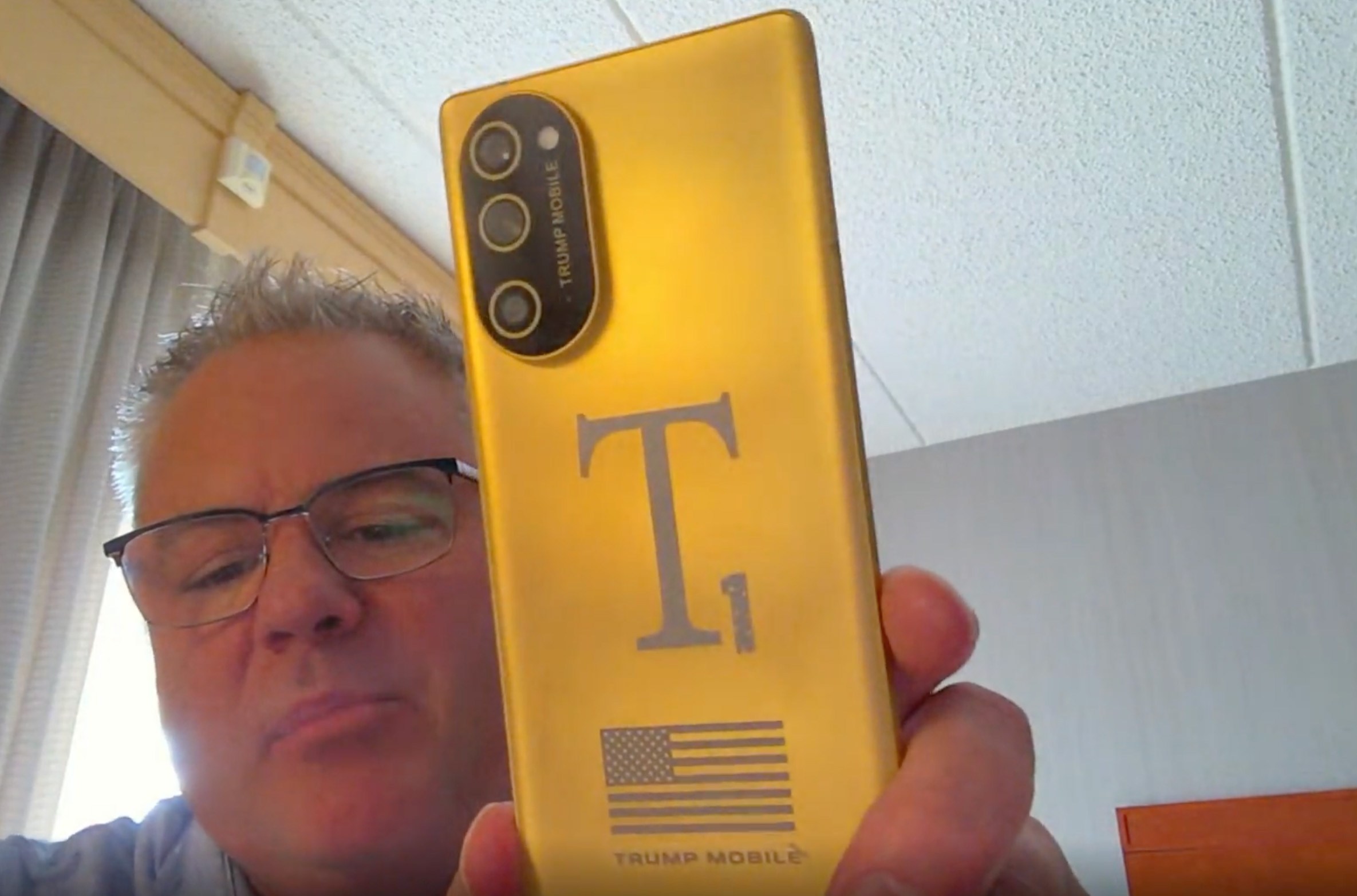 Screenshot of Eric Thomas from Trump Mobile holding a gold T1 Phone towards his webcam