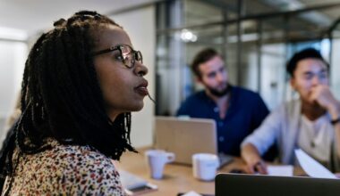 Three investors sit around a conference table with laptops and coffee cups while watching another investor present some data.