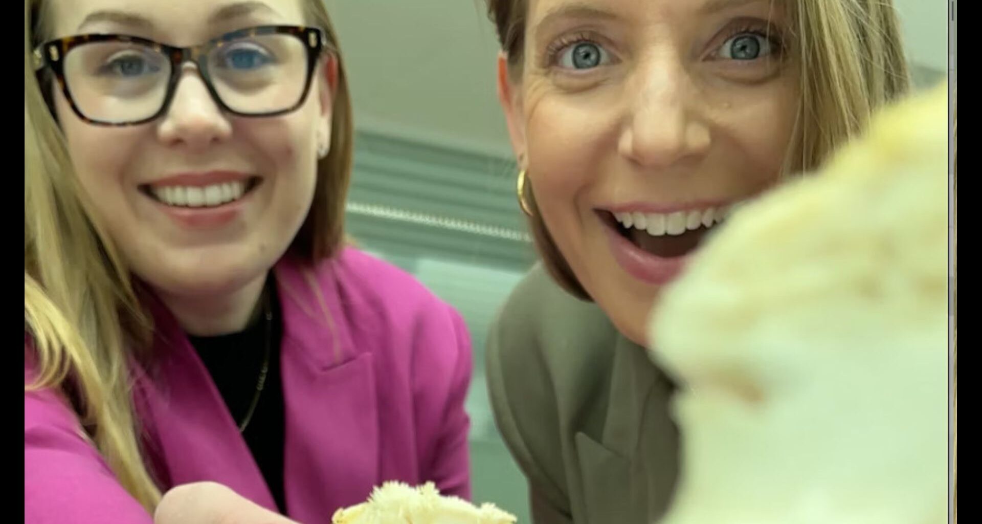 Two women hold up spongy white materials to the camera, one woman wears glasses and a pink blazer and the other has a gold hoop earring