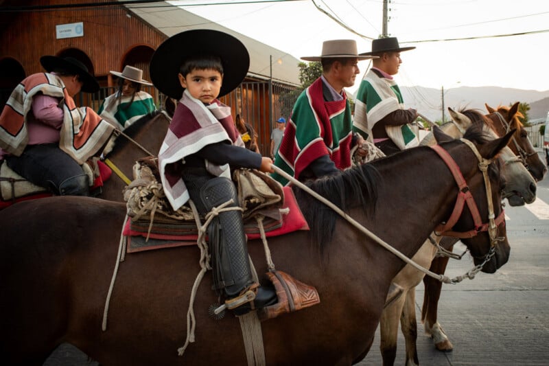 A young boy and several adults in traditional Chilean attire ride horses during a parade. The boy looks at the camera while others in ponchos and hats ride beside him on a street with mountains in the background.