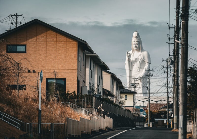 A large white statue of a serene figure stands behind residential houses, towering over the neighborhood with power lines crossing the scene under a cloudy sky.