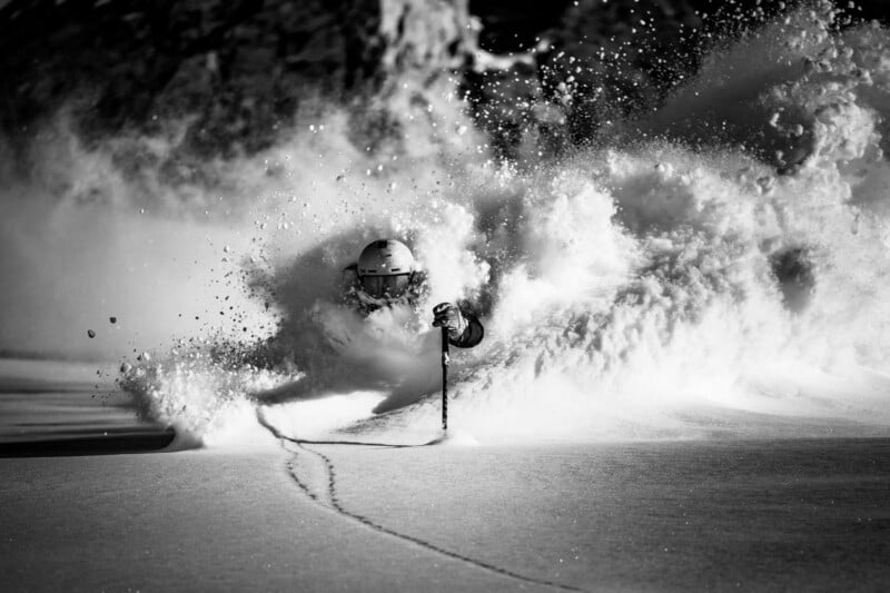 A skier rides through deep powder snow, creating a large spray that partially obscures their body. Only their helmet, goggles, and one ski pole are clearly visible. The scene is in black and white.